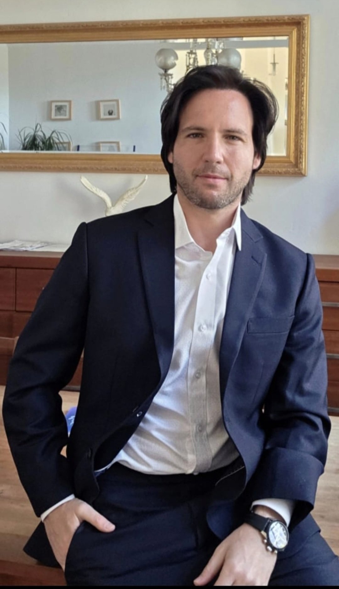 Man in navy blue suit and white shirt sitting indoors, with a framed mirror and home decor visible in the background